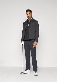 Conjunto de golfe masculino: casaco leve preto com detalhes cinzentos, polo preto, calças escuras, sapatos brancos com detalhes azuis e taco de golfe.