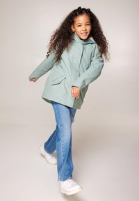 Giacca impermeabile verde menta con cappuccio, chiusura a bottoni e tasche laterali, abbinata a jeans azzurri e sneakers bianche.