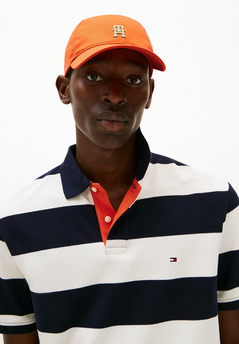 Polo a righe bianco e blu navy con colletto arancione. Cappellino arancione brillante con dettaglio del logo. Vestibilità regolare, tessuto testurizzato.