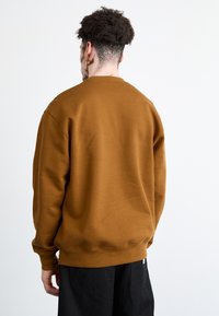 Sudadera marrón de material suave y texturizado, con cuello redondo, mangas largas y puños acanalados. Los pantalones negros son parcialmente visibles.