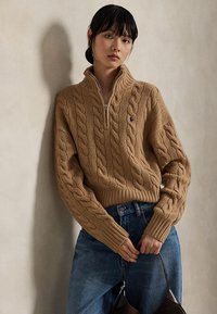 Femme portant un pull zippé en maille torsadée beige et un jean bleu, tenant un sac à main marron foncé sur un fond neutre.