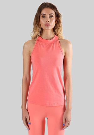 Femme aux cheveux longs portant un haut corail sans manches à col halter et un pantalon corail assorti, debout devant un fond gris clair uni.