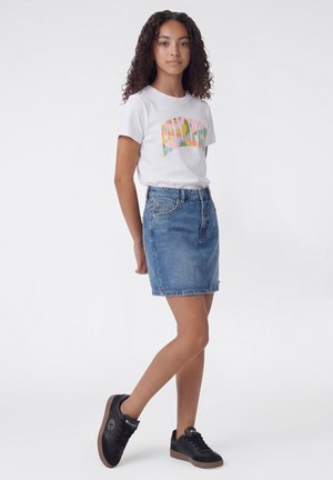 Junge Person mit lockigem Haar steht vor einem einfachen Hintergrund und trägt ein weißes T-Shirt mit Grafik, einen blauen Jeansrock und schwarze Turnschuhe.