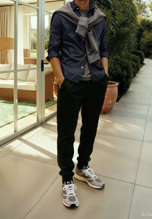 Man standing on tiled patio with hands in pockets, wearing dark shirt, black pants, sneakers, and a sweater draped over shoulders.