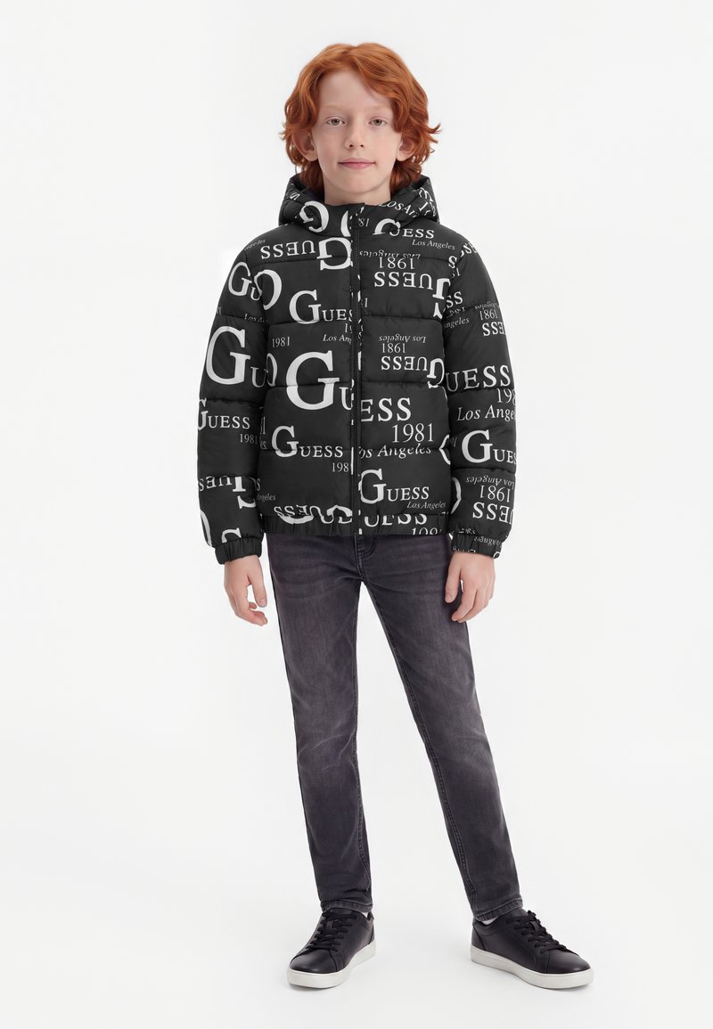 Enfant aux cheveux roux portant une doudoune noire avec le texte blanc GUESS, un jean foncé et des baskets noires, debout sur un fond blanc.