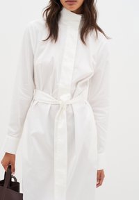 Weißes Hemdkleid mit hohem Kragen, langen Ärmeln und tailliertem Bund. Aus glattem Stoff mit subtiler Textur, hat es ein minimalistisches Design.
