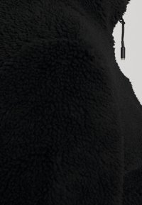 Zwart fleecejack met een zachte textuur, ritsluiting en een klein koord met een togglesluiting aan de linkerkant.