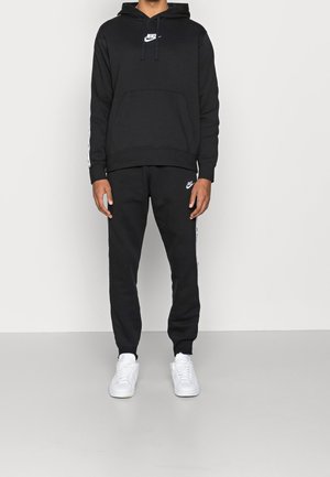 Homme portant un sweat à capuche noir Nike et un jogging assorti avec des baskets blanches, debout contre un mur uni.