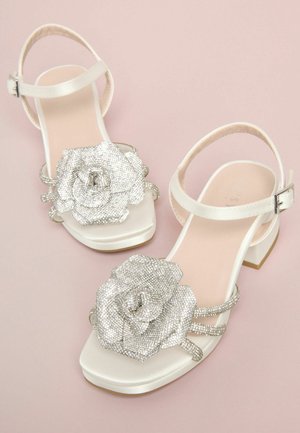 Weiße Sandale mit quadratischer Spitze, verziert mit einer großen silbernen Strassblume und mehreren dünnen Riemen. Niedriger Blockabsatz.