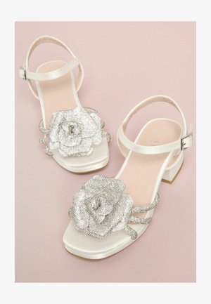 Weiße Sandale mit quadratischer Spitze, verziert mit einer großen silbernen Strassblume und mehreren dünnen Riemen. Niedriger Blockabsatz.
