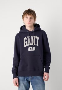 Marineblå hettegenser laget av bomull med en frontlomme, som har et hvitt grafisk trykk av "GANT 49." Designet inkluderer snordetaljer.