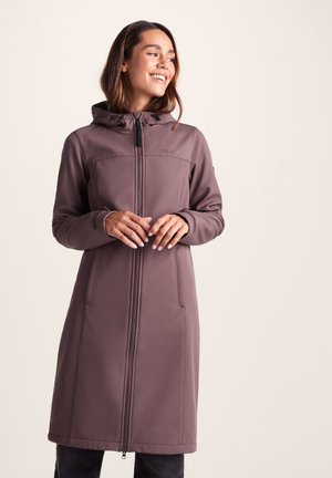 Woman wearing a long mauve hooded coat with a front zipper, standing against a plain light background, smiling and looking to the side.