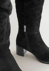Juodos suede aukštakulnės botas iki kelių turi keturkampį kulną ir šoninės užtrauktuko užsegimą. Medžiaga yra minkšta, su matomomis siūlėmis palei siūles.