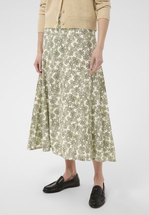 Person trägt eine beige Strickjacke, weißen Rock mit grünem Blumenmuster und schwarze Loafer und steht vor einem einfarbigen Hintergrund.