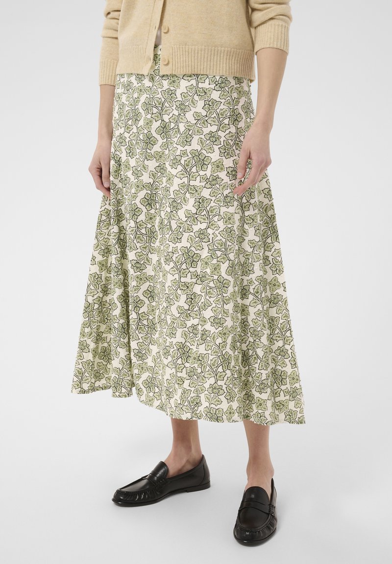 Personne portant un cardigan beige, une jupe blanche avec un imprimé floral vert et des mocassins noirs, debout devant un fond uni.