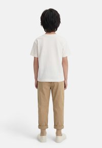 Παιδί που στέκεται με την πλάτη γυρισμένη, φοράει λευκό πουκάμισο με κοντά μανίκια, μπεζ παντελόνι γυρισμένο προς τα πάνω, μπεζ κάλτσες και λευκά παπούτσια σε λευκό φόντο.