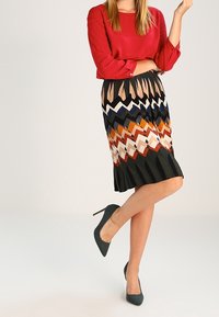 Blouse rouge à manches retroussées, associée à une jupe longueur genoux arborant un motif zigzag multicolore aux teintes terreuses. Talons hauts vert foncé.