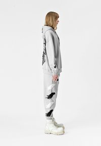 Sudadera gris de gran tamaño con bordado de alas negras, combinada con pantalones jogger grises que presentan patrones de alas en blanco y negro, y botas claras.