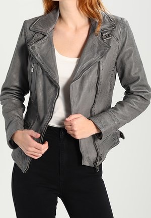 Femme portant une veste de motard en cuir grise avec des fermetures éclair et des boucles sur un top blanc et un pantalon noir, tenant les bords avant de la veste.