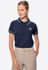 Polo navy con dettagli bianchi, colletto a tre bottoni e logo ricamato. Indossato con pantaloni beige e una cintura coordinata.