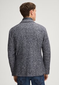 Blazer grigio con motivo a trama e tessuto testurizzato, colletto scuro. Presenta una singola spaccatura e polsini con bottoni, abbinato a jeans blu.