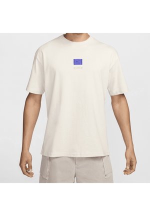 Homme portant un t-shirt beige clair uni avec un petit logo carré bleu sur la poitrine et un short beige clair avec des poches latérales.