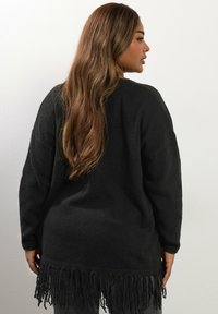 Kvinna med långt brunt hår som bär en svart fransad tröja, vänd mot höger framför en enkel vit vägg.