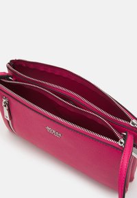 Vaaleanpunainen tekstuurinen käsilaukku, jossa on kaksi vetoketjullista osastoa ja hopeiset yksityiskohdat. Edessä on logo ja ohut, yhteensopiva olkahihna.