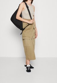 Vrouw in een beige cargomidirok, een mouwloze beige top, zwart-witte sneakers en met een grote zwarte schoudertas.