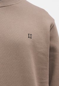 Ανοιχτό καφέ φούτερ με ριμπ στο λαιμό, που διαθέτει μικρό κεντητό σκούρο καφέ «II» στο στήθος. Λείο, υφασμάτινο ύφασμα με υφή.