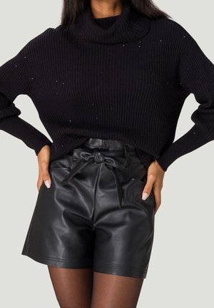 Femme portant un pull-col roulé noir à côtes avec de minuscules paillettes et un short en cuir noir taille haute noué avec une ceinture en nœud.
