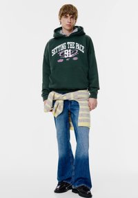 Felpa con cappuccio verde scuro con scritte bianche e rosa, jeans blu a zampa di elefante e un maglione a righe pastello annodato in vita. Scarpe nere.