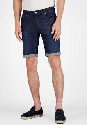 Man draagt donkerblauwe opgerolde denim shorts en zwarte instapschoenen, staand tegen een witte achtergrond.