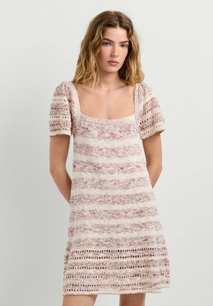 Blonde Frau trägt ein kurzärmliges, gestricktes Kleid mit rosa und cremefarbenen horizontalen Streifen und steht vor einem einfarbigen hellen Hintergrund.