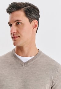 Jasno beżowy sweter v-neck wykonany z miękkiego dzianinowego materiału, z prążkowanym kołnierzem i delikatną fakturą. Nosi się go na białej koszulce.