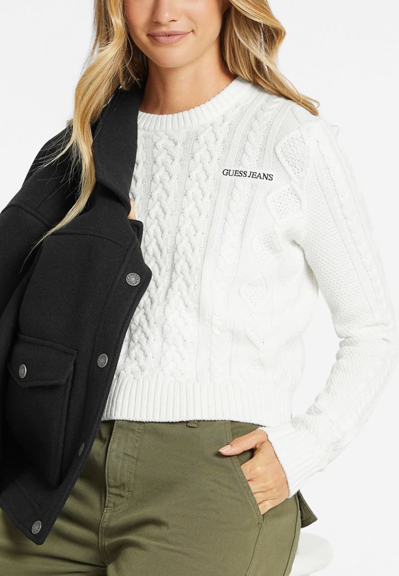 Weißer Strickpullover mit Zopfmuster, gerippten Bündchen und Saum, mit dem Logo "GUESS JEANS". Kombiniert mit einer schwarzen Jacke und olivgrünen Hosen.