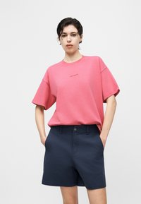 Άτομο με κοντά σκουρόχρωμα μαλλιά, φοράει ροζ φαρδιά μπλουζά T-shirt και σκούρο μπλε σορτς, στέκεται με τα χέρια στις τσέπες μπροστά σε απλό φόντο.