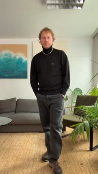 Melns augsts apkakles džemperis, pelēkas džinsu bikses, brūnas kurpes. Iekštelpu vide ar dīvānu, stikla galdu un augu. Sienas māksla attēlo zilas viļņus.