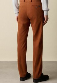 Hnědé bavlněné kalhoty se střihovým designem rovného nohavice, s kapsami na zadní straně a jemnými detaily stehu. Nosené s černými botami.
