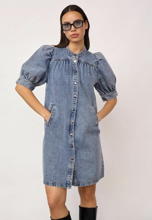 Donna che indossa un vestito di jeans al ginocchio con maniche a sbuffo e bottoni sul davanti, mani nelle tasche e stivali neri.