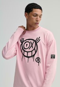 Hosszú ujjú rózsaszín pamut póló, elöl egy nagy fekete grafikával és arccal a dizájnon, valamint egy kis márkacímkével az ujjon.