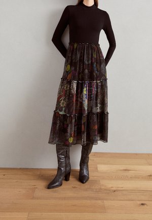 Femme portant un haut noir à manches longues, une jupe en voile multi-couches avec des motifs colorés et des bottes métalliques texturées montant jusqu'aux genoux, debout sur un sol en bois.