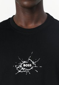 Fekete pamut póló, fehér grafikával, amelyen egy repedezett felület látható, benne a "BOSS" felirattal. Kerek nyakkivágású kialakítás.