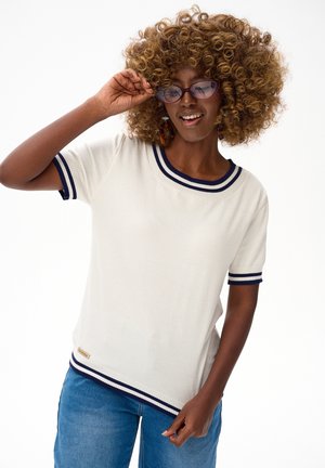 Kobieta o kręconych włosach, w okularach, ubrana w biały sweter z krótkim rękawem i granatowymi paskami oraz niebieskie dżinsy, uśmiechająca się na białym tle.