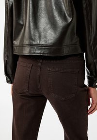 Giubbotto di pelle nera sopra jeans di denim marrone scuro, che mostrano le tasche posteriori e una texture liscia con sottili dettagli di cucitura.
