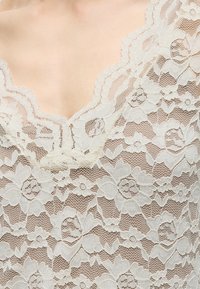 Gros plan d’un tissu en dentelle florale blanche sur la peau montrant des motifs détaillés de fleurs et de feuilles avec des bords festonnés.