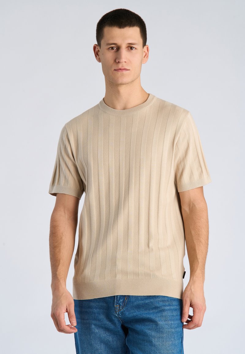 Junger Mann, der ein beiges, kurzärmliges, geripptes Strickshirt und blaue Jeans trägt, steht vor einem einfarbigen hellen Hintergrund.