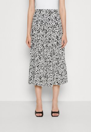 Femme portant une jupe midi fleurie noire et blanche à taille haute et des talons ouverts noirs, debout devant un fond uni.