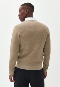 Jersey de punto beige con un acabado texturizado, escote redondo y mangas largas. Superpuesto a una camisa blanca con cuello. Pantalones negros visibles.