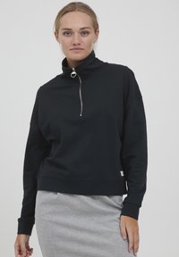 Black zip-up sweatshirt with a high collar, featuring a chevron design and ribbed cuffs. Soft fabric, cropped fit. Grey skirt visible below.